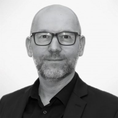 Marcel Kamin Mann mit Brille, kurzem Haarschnitt und Bart, trägt einen schwarzen Anzug.